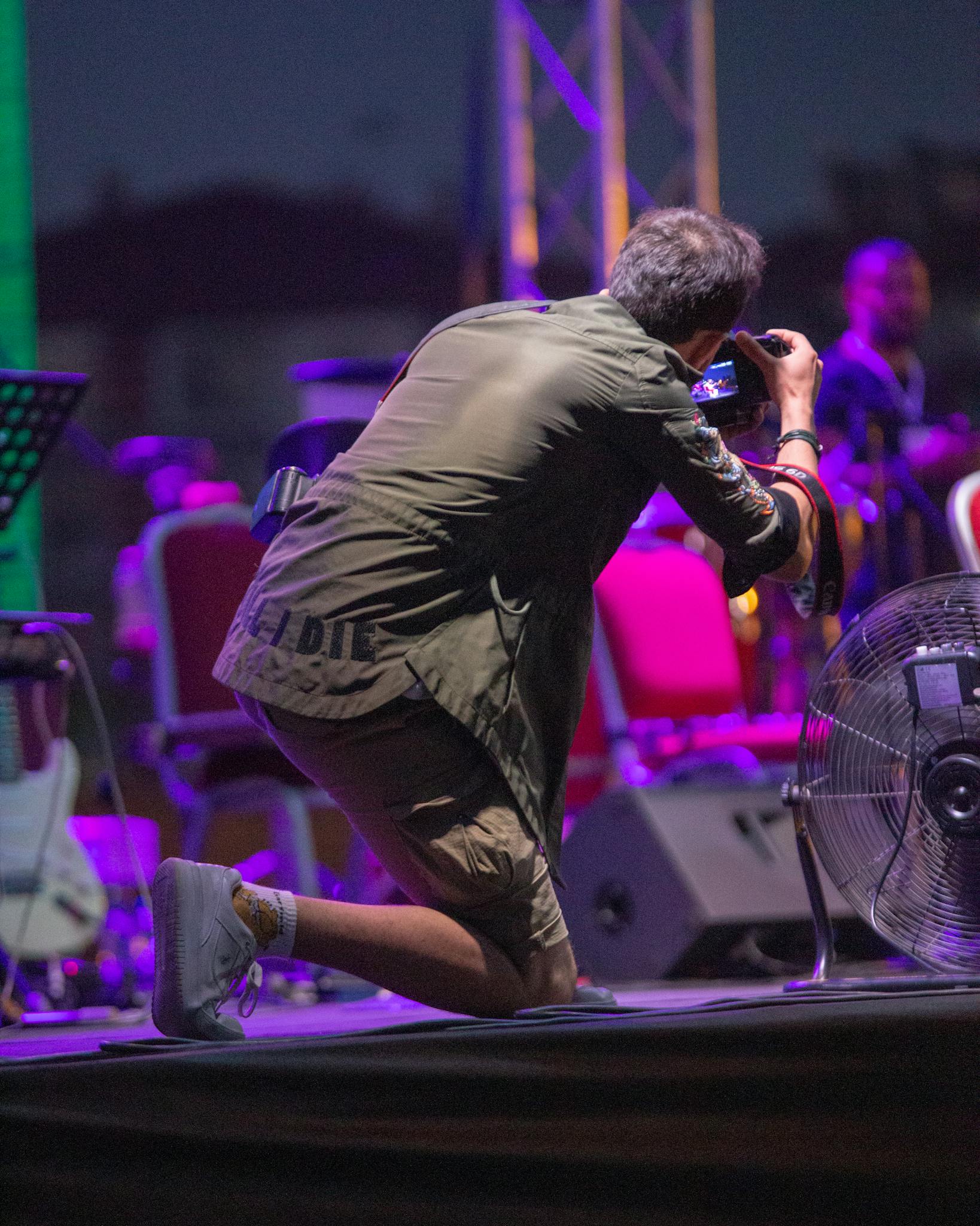 A photographer kneels on stage capturing a vibrant concert scene at night.